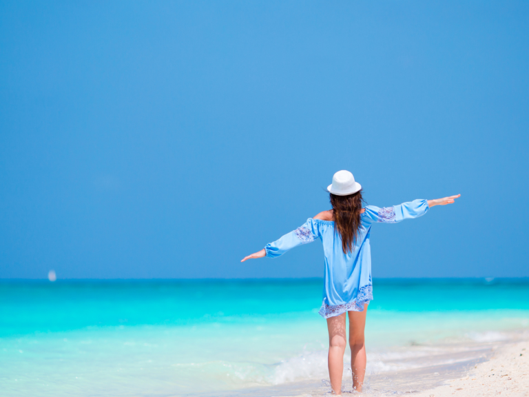 Woman in blue top standing on a white beach, with arms raised signaling freedom after therapy in palm beach gardens with Erin Pallard.